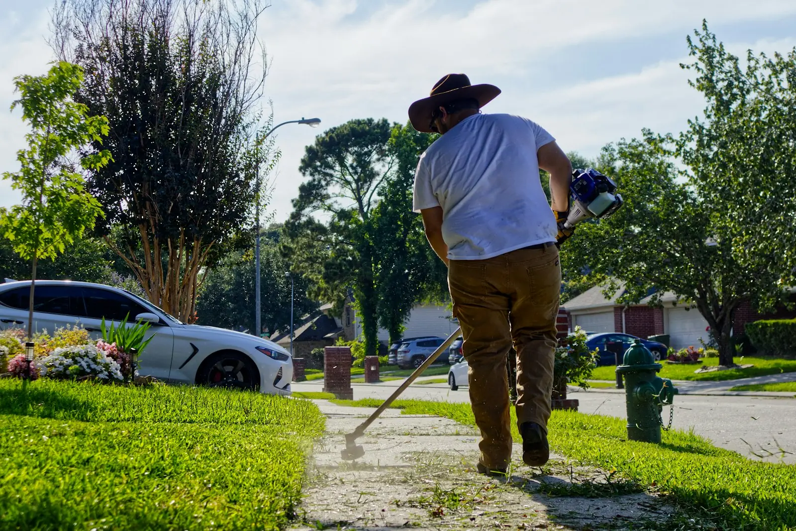 Landscaper in a wide-brim hat edging grass along a suburban footpath